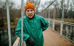 An older man in a green rain jacket leans on the side of a bridge after jogging.