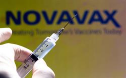 Gloved fingers hold a half-full syringe in front of a Novavax company sign.