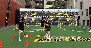 Four people in soccer gear on a mini soccer pitch in an urban environment, text on the pitch reads No Pain More Gain