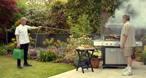 Gordon Ramsay holds an Uber Eats bag while spraying a grill with water from a hose as the man working the grill looks on