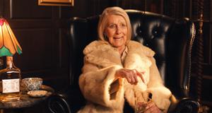 A woman wearing a fur coat and holding a glass of tequila sits in a leather chair