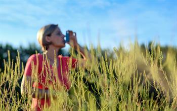 A woman in an orange T-shirt stands in a field against a blue sky, taking a puff from her metered dose inhaler
