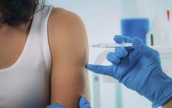 A woman wearing a white vest as a blue-gloved hand of a healthcare professional are seen injecting a vaccine.