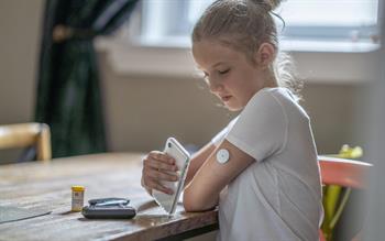 A girl in a white T-shirt reads her blood glucose level off a smartphone held next to a sensor on her arm.