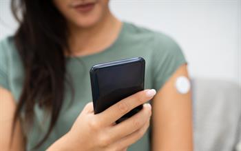 A young woman with a glucose monitoring patch on her arm consults her blood glucose reading on her smartphone