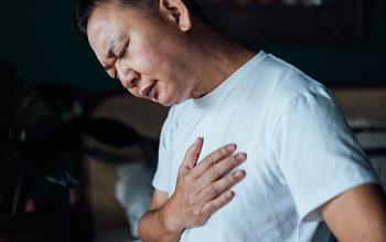 Close up of an Asian man holding his chest in discomfort, suffering from chest pain while sitting on bed at home.