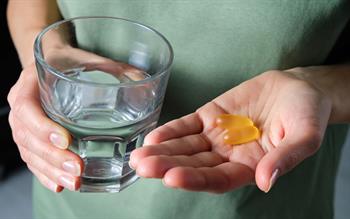 A woman in a pale green top holds two yellow soft capsules in one hand and a glass of water in the other.