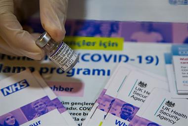 A close up on a vial of COVID-19 vaccine with promotional material about the vaccination programme in the background