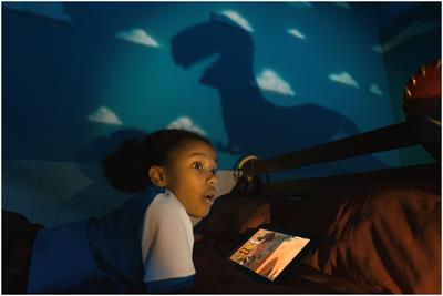Child on bed watching Toy Story 3 on their iPad with a shadow of a dinosaur on the bedroom wall