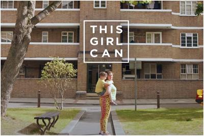 Women stands smiling in front of a block of flats, with a child on her back 