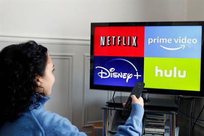 Logos of media service providers, Netflix, Amazon Prime Video, Disney + and Hulu are displayed on the screen of a television as a woman holds up a TV remote