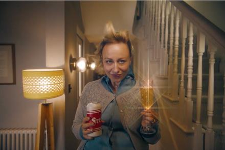 A woman stands in her hallway, staring somewhat menacingly at the camera while holding a can of lager in one hand and a champagne glass of beer in the other