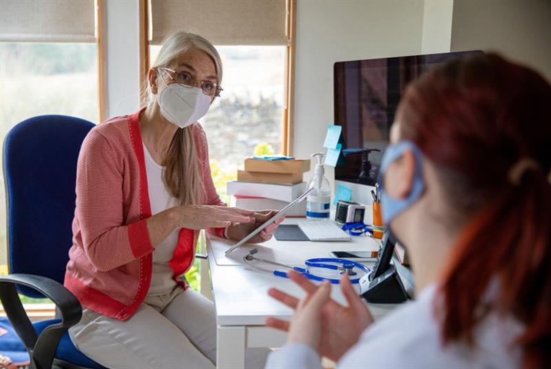 GP wearing surgical mask speaks to patient in consulting room