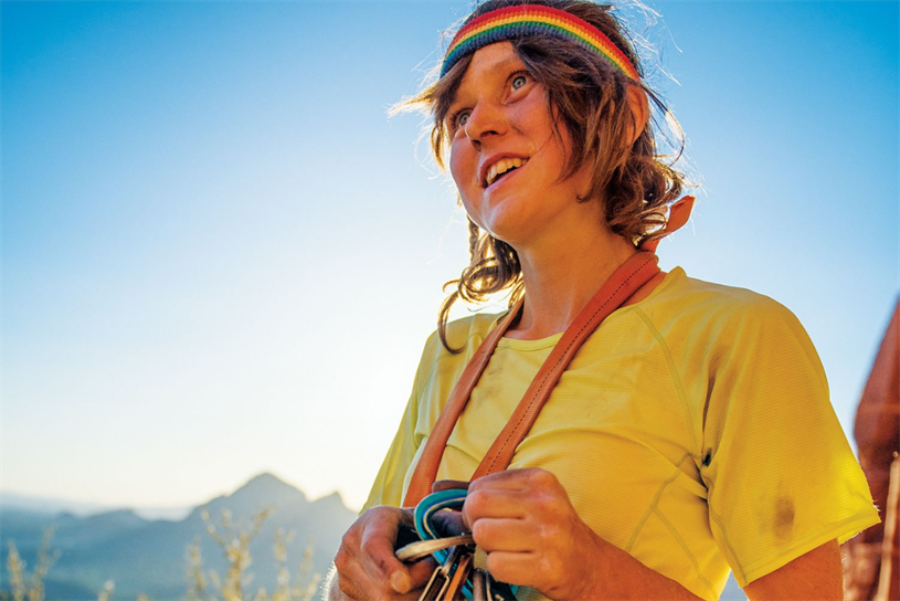 An image of climber Lor Sabourin standing in the mountains, wearing a bright yellow t-shirt and a rainbow-hued sweatband