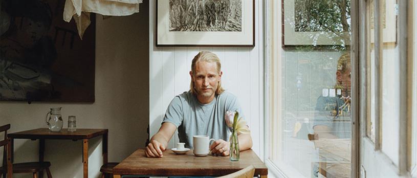 Image of Felix Richter sitting at a cafe table by a window with a mug on the table in front of him