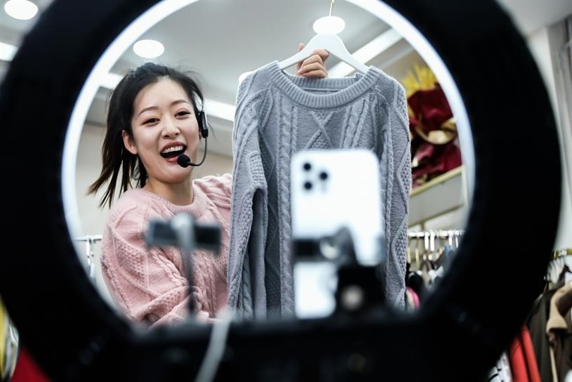 Smiling Asian woman holding sweater in a department store on shopping livestream.