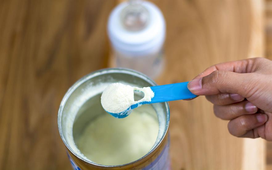 A woman's hand holds a blue plastic scoop full of baby milk powder above the tin she has just filled it from, with a baby bottle visible in the background.