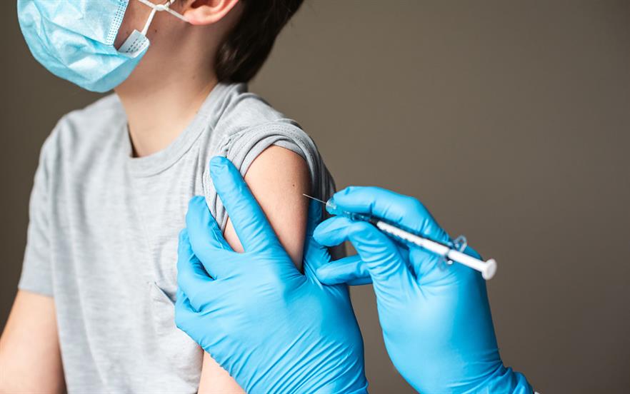 A young boy wearing a face mask looks away as a doctor's blue-gloved hands are seen injecting a vaccine into the boy's upper arm.