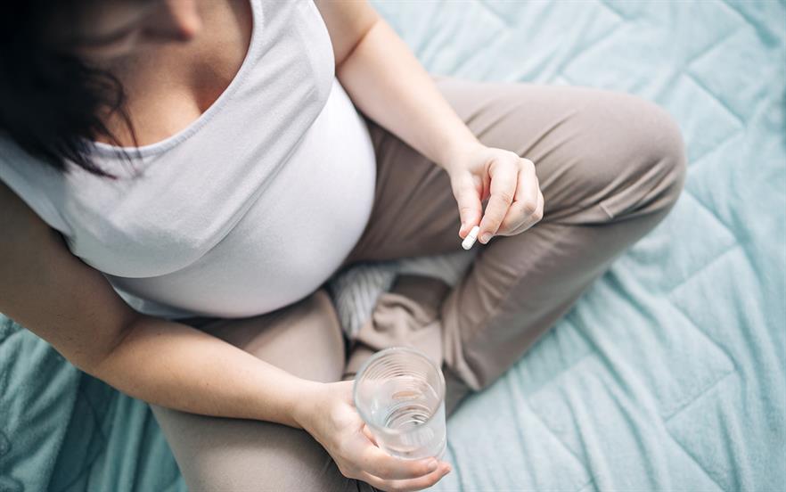 A pregnant woman is seen from above as she sits cross-legged on a bed taking a pill with a glass of water.