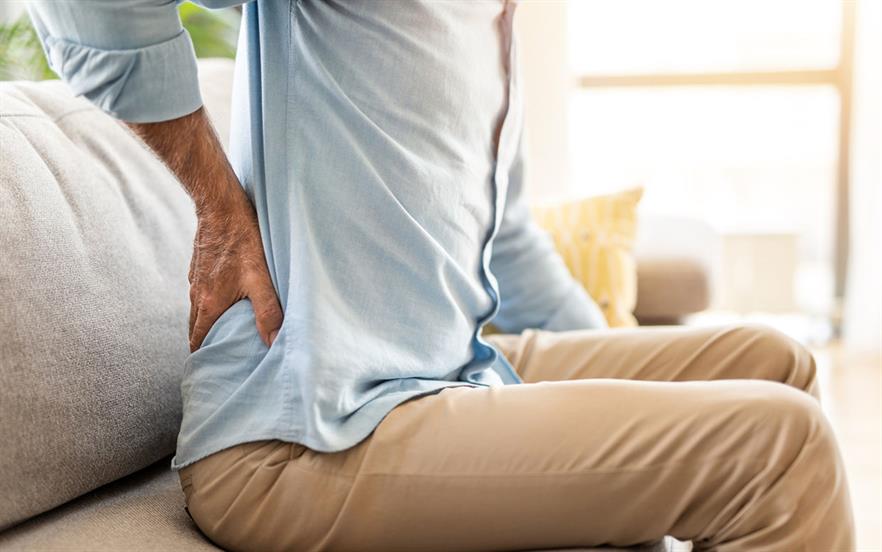 An older man in a blue shirt and beige trousers leans forward on a sofa rubbing his painful lower back.