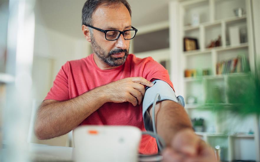 A middle-aged man with glasses and a beard checks his blood pressure at home.