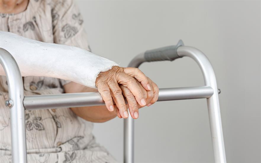 An elderly woman with one arm in a plaster cast sits resting both hands on her walker